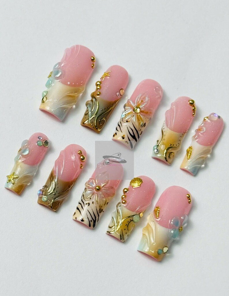 May include: A set of ten artificial nails with a pink and white gradient design. Each nail features intricate embellishments, including gold accents, small beads, and floral or seashell-inspired decorations. The nails have a glossy finish.