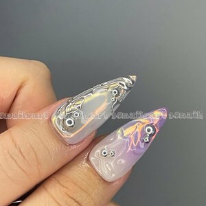 May include: Close-up of a hand with long, pointed nails. The nails are painted with a light purple and white ombre effect and have silver accents. The nails are decorated with small, silver beads.