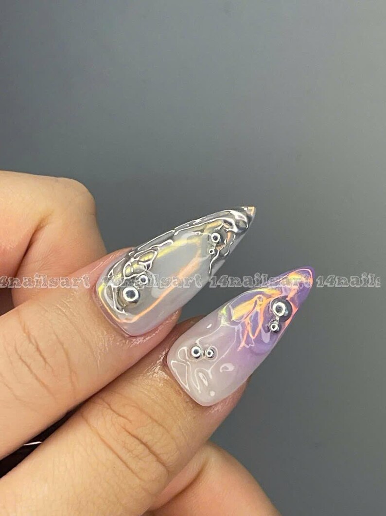 May include: Close-up of a hand with long, pointed nails. The nails are painted with a light purple and white ombre effect and have silver accents. The nails are decorated with small, silver beads.
