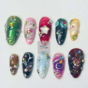 Pode incluir: Um conjunto de dez unhas artificiais com vários designs. As unhas apresentam uma gama de cores, incluindo verde, rosa, vermelho, preto e amarelo. Cada unha é adornada com embelezamentos como strass, pérolas e detalhes em forma de estrela, criando um visual glamouroso.
