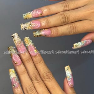 May include: Long, square-shaped artificial nails with a pink base, adorned with gold and white floral designs, rhinestones, and zebra print accents. The text "14nailsart" is visible on some nails. A stylish manicure.