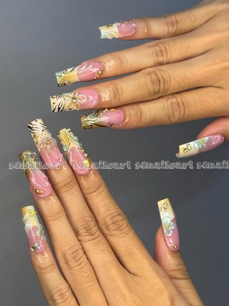 May include: Long, square-shaped artificial nails with a pink base, adorned with gold and white floral designs, rhinestones, and zebra print accents. The text "14nailsart" is visible on some nails. A stylish manicure.