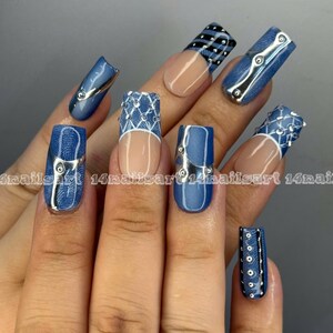 May include: A hand displaying a set of long, artificial nails with a denim-inspired design. The nails feature a mix of blue denim patterns, silver accents, and clear sections. Some nails have a silver metallic trim.