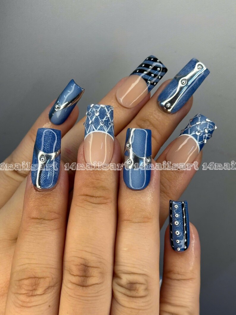 May include: A hand displaying a set of long, artificial nails with a denim-inspired design. The nails feature a mix of blue denim patterns, silver accents, and clear sections. Some nails have a silver metallic trim.