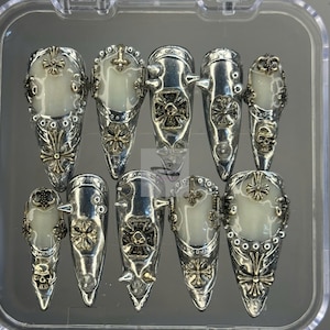 Puede incluir: Un conjunto de diez cubiertas de uñas puntiagudas de color plateado con diseños ornamentados de inspiración gótica. Cada uña presenta un óvalo central blanquecino, rodeado de intrincados trabajos en metal, incluyendo cruces, patrones florales y detalles de calaveras. Las uñas se muestran en una caja de plástico transparente.