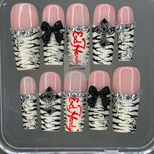 May include: A set of ten artificial nails with a pink and white zebra print design. Some nails feature black bows, silver crosses, or the text "Ed Hardy" in red. Each nail has a pink tip and is decorated with silver rhinestones.