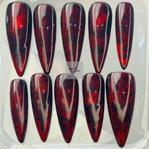 Pode incluir: Um conjunto de dez unhas postiças longas em forma de estilete. Cada unha apresenta uma base vermelha escura brilhante com um efeito marmorizado de preto e tons de vermelho mais claros. As unhas estão dispostas em um recipiente de plástico transparente.