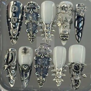 May include: A set of ten long, pointed false nails. The nails feature a variety of designs, including silver embellishments, spikes, skulls, crosses, and lace patterns. Some nails have a solid white base, while others are silver or dark blue.