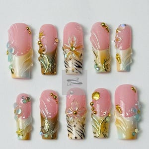 May include: A set of ten artificial nails with a pink and white color scheme, adorned with gold accents, seashells, and floral designs. The nails feature a glossy finish and various embellishments, creating a detailed and artistic appearance.