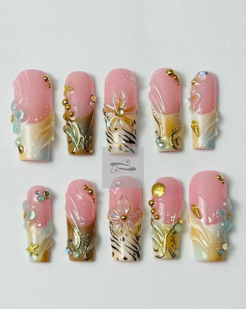 May include: A set of ten artificial nails with a pink and white color scheme, adorned with gold accents, seashells, and floral designs. The nails feature a glossy finish and various embellishments, creating a detailed and artistic appearance.