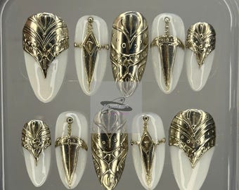 May include: A set of ten press-on nails in a clear plastic case. Each nail is painted white with gold-colored embellishments. The embellishments include ornate designs and sword-like accents.