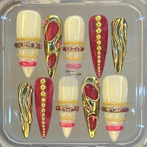 Puede incluir: Un conjunto de diez uñas artificiales en una caja de plástico transparente. Las uñas presentan una variedad de diseños, incluyendo detalles dorados, purpurina roja y cadenas doradas. Algunas uñas son de color crema sólido, mientras que otras son de color rojo intenso. Las uñas están diseñadas para la moda y la belleza.