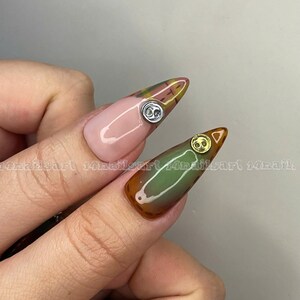 K&ouml;nnte beinhalten: Nahaufnahme von zwei langen, mandelf&ouml;rmigen N&auml;geln mit einem gr&uuml;n-braunen Ombre-Design. Ein Nagel hat einen silbernen Knopf als Verzierung, der andere einen goldenen Knopf.