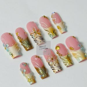 May include: A set of ten artificial nails with a pink and white design. Each nail features a pink base with a white tip, adorned with gold, clear, and iridescent embellishments, including flowers, shells, and gems. The nails have a glossy finish.