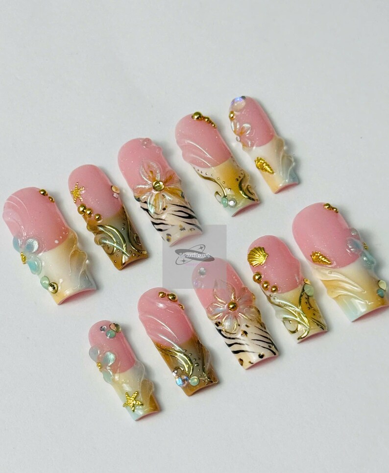 May include: A set of ten artificial nails with a pink and white design. Each nail features a pink base with a white tip, adorned with gold, clear, and iridescent embellishments, including flowers, shells, and gems. The nails have a glossy finish.