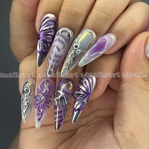 May include: Purple and silver butterfly nail art with a clear iridescent base. The nails are long and pointed.
