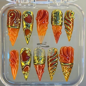 Peut inclure: Un ensemble de dix faux ongles dans un étui en plastique transparent. Les ongles présentent un dégradé d'orange à rouge, avec des accents décoratifs dorés et bruns. Le texte "14nailsart" est visible sur l'étui.