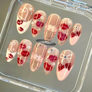 Pode incluir: Um conjunto de dez unhas artificiais com v&aacute;rios designs. As unhas apresentam uma base nude com l&aacute;bios vermelhos, cora&ccedil;&otilde;es e padr&otilde;es de renda. Algumas unhas t&ecirc;m strass e detalhes brancos. O texto "14 nailsart" &eacute; vis&iacute;vel.