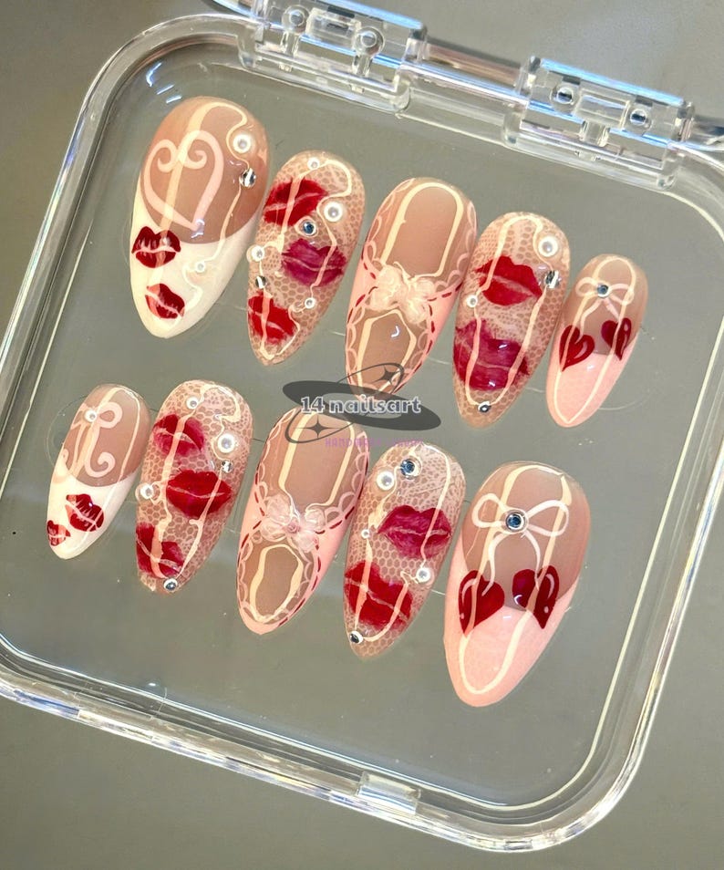 Pode incluir: Um conjunto de dez unhas artificiais com v&aacute;rios designs. As unhas apresentam uma base nude com l&aacute;bios vermelhos, cora&ccedil;&otilde;es e padr&otilde;es de renda. Algumas unhas t&ecirc;m strass e detalhes brancos. O texto "14 nailsart" &eacute; vis&iacute;vel.