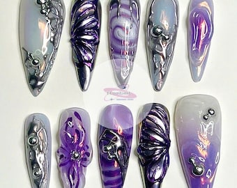 Puede incluir: Conjunto de diez uñas largas, puntiagudas, iridiscentes de color púrpura y plateado con varios diseños, incluyendo mariposas, remolinos y flores.