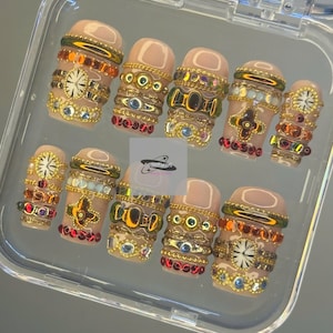 May include: A collection of ten artificial nails presented in a clear case. Each nail features a nude base and is decorated with gold-tone embellishments, such as rhinestones, beads, and watch-face motifs. The nails are arranged in a row, highlighting the detailed craftsmanship.