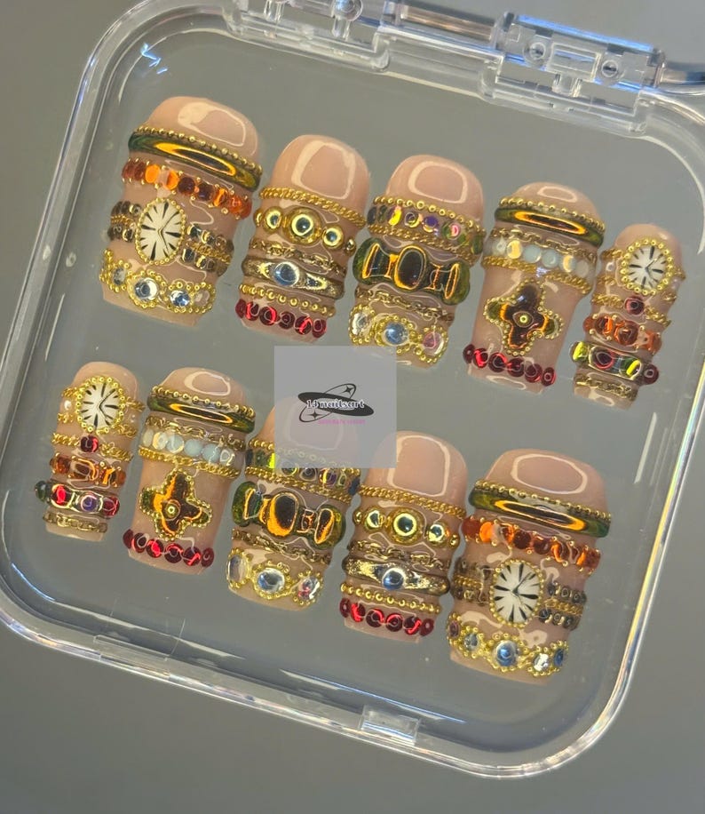 May include: A collection of ten artificial nails presented in a clear case. Each nail features a nude base and is decorated with gold-tone embellishments, such as rhinestones, beads, and watch-face motifs. The nails are arranged in a row, highlighting the detailed craftsmanship.