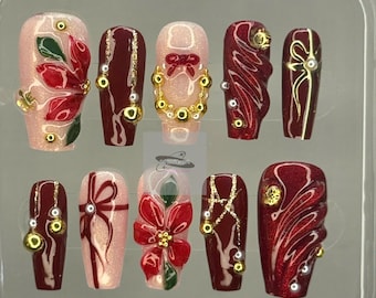 May include: A set of ten artificial nails in a clear plastic case. The nails feature various designs, including red and gold accents, bows, and floral patterns. Some nails have gold and pearl embellishments. The nails are in a variety of shapes.