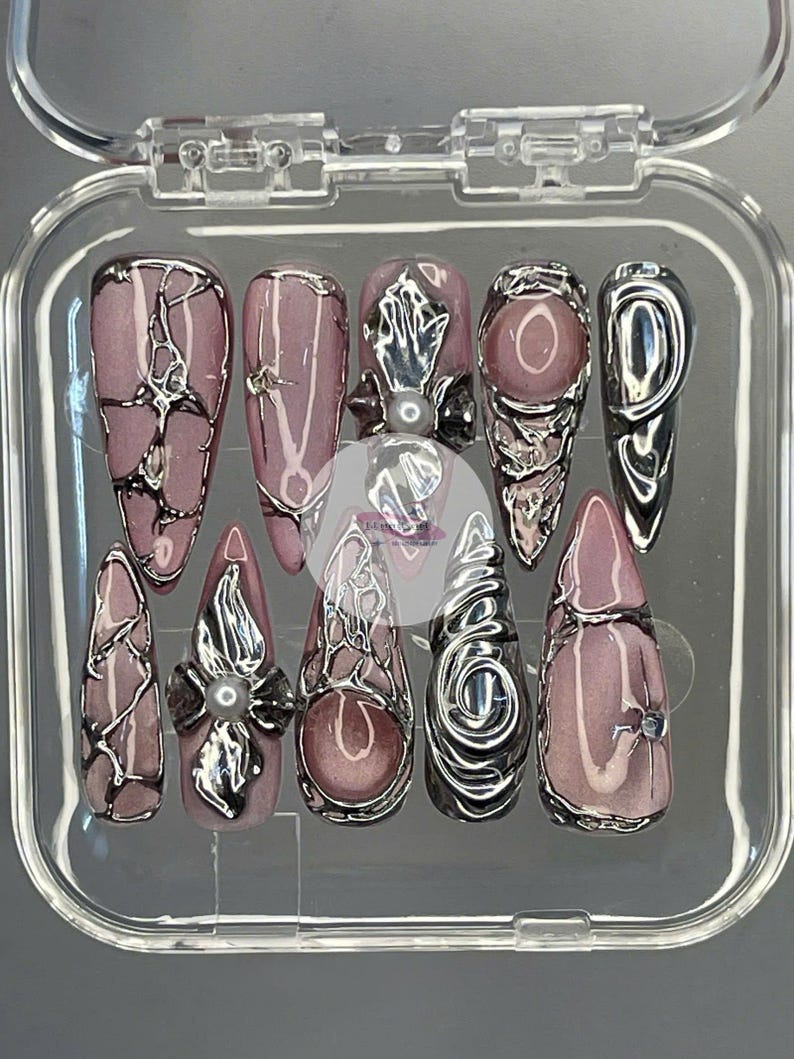May include: A set of ten artificial nails in a clear plastic case. The nails are primarily mauve with silver metallic accents. Some nails feature intricate silver designs, while others have small embellishments.