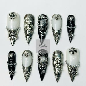 May include: A collection of ten long, pointed false nails. The nails feature a variety of designs, including white and black bases, silver metal accents, crosses, and decorative embellishments. The nails are arranged on a white surface.