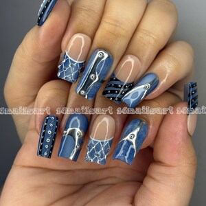 May include: A hand displaying a set of long, square-shaped artificial nails. The nails feature a denim-inspired design with silver accents, including rivets and lace-like patterns. Some nails have a clear base with a white tip.