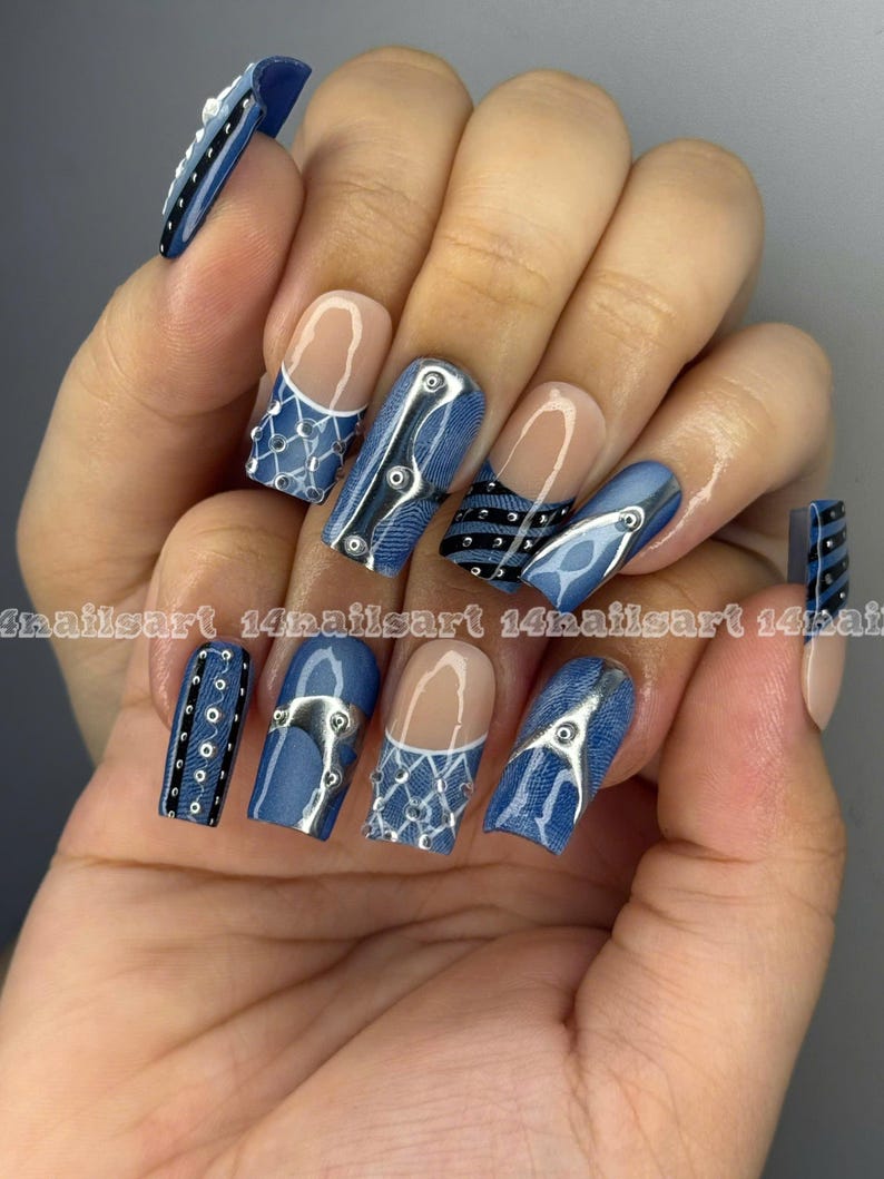 May include: A hand displaying a set of long, square-shaped artificial nails. The nails feature a denim-inspired design with silver accents, including rivets and lace-like patterns. Some nails have a clear base with a white tip.