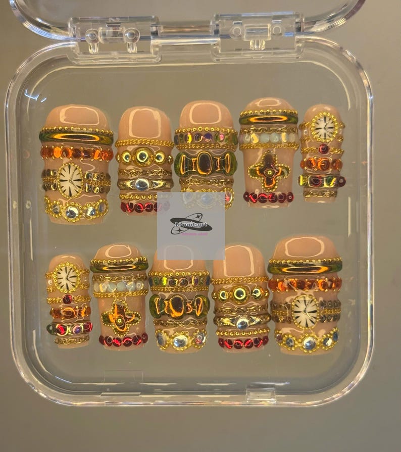 May include: A set of ten artificial nails in a clear plastic case. The nails are a nude colour and decorated with gold, red, and clear rhinestones. The designs include floral and geometric patterns.