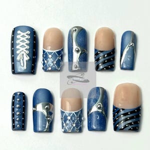 May include: A set of ten artificial nails with various designs. The nails feature a mix of blue denim, silver accents, and nude-colored tips. Some nails have a lace-up design, while others have silver embellishments or striped patterns.