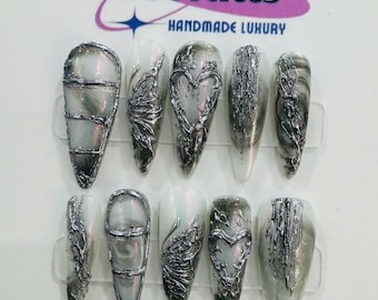 Puede incluir: Conjunto de diez uñas postizas blancas con purpurina plateada y diseños metálicos. Los diseños incluyen corazones, alas y remolinos. Las uñas están dispuestas en dos filas sobre una superficie blanca. El texto "14 Nails Handmade Luxury" es visible en la parte superior de la imagen.