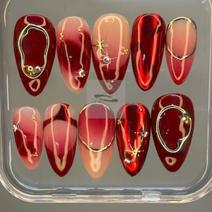 May include: A collection of ten artificial nails displayed in a clear case. The nails showcase a glossy red and pink gradient, adorned with gold-coloured accents and rhinestones. Some nails feature gold outlines or starburst patterns.