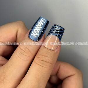 May include: Close-up of two fingernails with nail art. One nail features a blue background with white shoelace design and black borders. The other nail has a blue and black striped design with a nude base and silver studs.