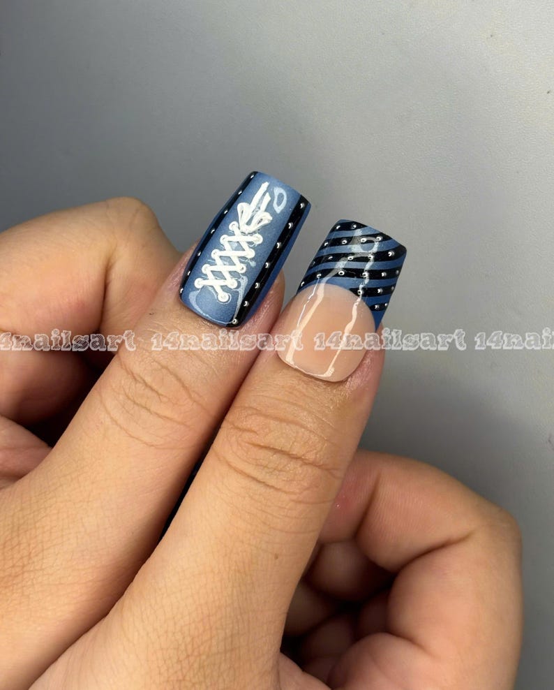 May include: Close-up of two fingernails with nail art. One nail features a blue background with white shoelace design and black borders. The other nail has a blue and black striped design with a nude base and silver studs.