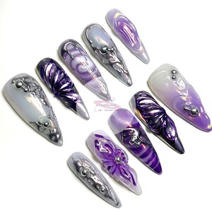 May include: Set of ten long, pointed, purple and silver nail tips with various designs. Some nails have a swirl design, others have a 3D flower design, and some have a silver glitter design.