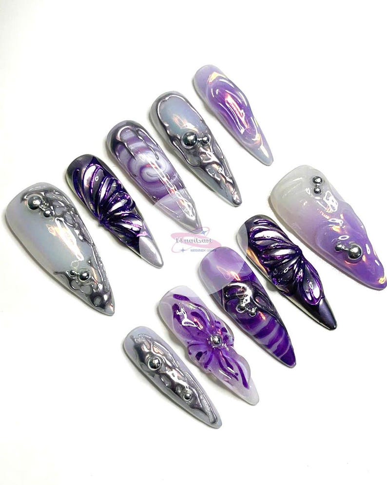 May include: Set of ten long, pointed, purple and silver nail tips with various designs. Some nails have a swirl design, others have a 3D flower design, and some have a silver glitter design.