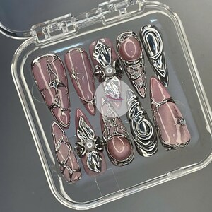 May include: A set of ten artificial nails in a clear plastic case. The nails are mauve with silver metallic accents. Some nails feature a pearl embellishment, and others have a rhinestone.
