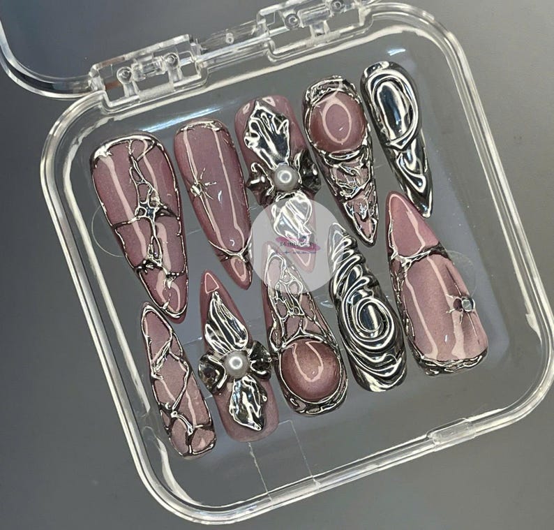 May include: A set of ten artificial nails in a clear plastic case. The nails are mauve with silver metallic accents. Some nails feature a pearl embellishment, and others have a rhinestone.