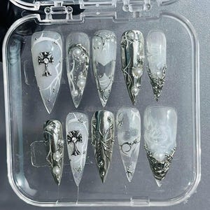 May include: A set of ten white press-on nails with silver accents. The nails feature a variety of designs, including crosses, butterflies, and vines. The nails are displayed in a clear plastic case.