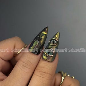 Peut inclure: Deux ongles longs et pointus, vert et or iris&eacute;s, avec un motif textur&eacute;. Un ongle pr&eacute;sente deux petits motifs d'yeux d&eacute;taill&eacute;s. Les ongles sont orn&eacute;s de bagues en or.