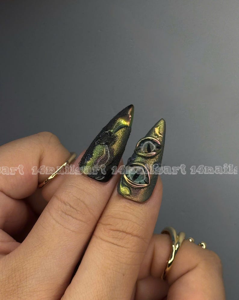 Peut inclure: Deux ongles longs et pointus, vert et or iris&eacute;s, avec un motif textur&eacute;. Un ongle pr&eacute;sente deux petits motifs d'yeux d&eacute;taill&eacute;s. Les ongles sont orn&eacute;s de bagues en or.