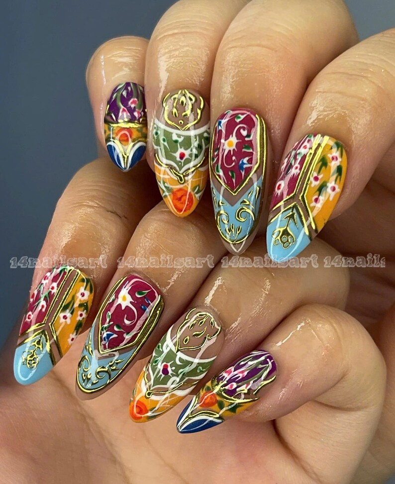 May include: A hand with long, almond-shaped nails, each adorned with intricate designs. The nails feature a gold outline with colorful patterns in shades of blue, orange, green, and purple. The text "14nailsart 14nailsart 14nails" is visible.