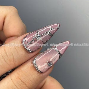 May include: Close-up of two long, almond-shaped nails with a light pink base and silver wire-like designs. One nail features a small, sparkling gem. The nails are part of a manicure, showcasing nail art.