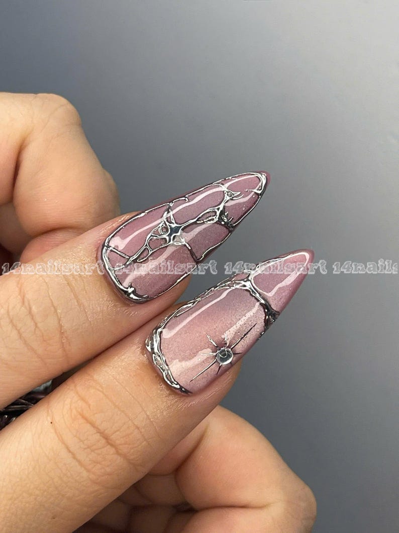 May include: Close-up of two long, almond-shaped nails with a light pink base and silver wire-like designs. One nail features a small, sparkling gem. The nails are part of a manicure, showcasing nail art.
