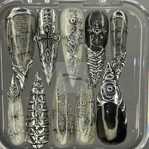 Puede incluir: Un conjunto de diez uñas postizas largas y puntiagudas en una caja de plástico transparente. Las uñas presentan una variedad de diseños, que incluyen adornos de metal plateado, cruces y patrones de escritura en tonos grises y negros.