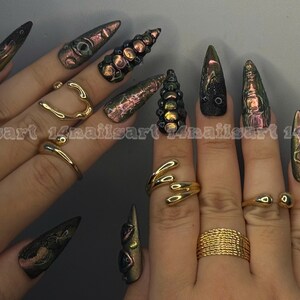 Peut inclure: Mains avec de longs ongles pointus iris&eacute;s verts, violets et dor&eacute;s. Les doigts sont orn&eacute;s de bagues dor&eacute;es de diff&eacute;rents mod&egrave;les, dont un c&oelig;ur, une forme enroul&eacute;e et une bande tress&eacute;e.