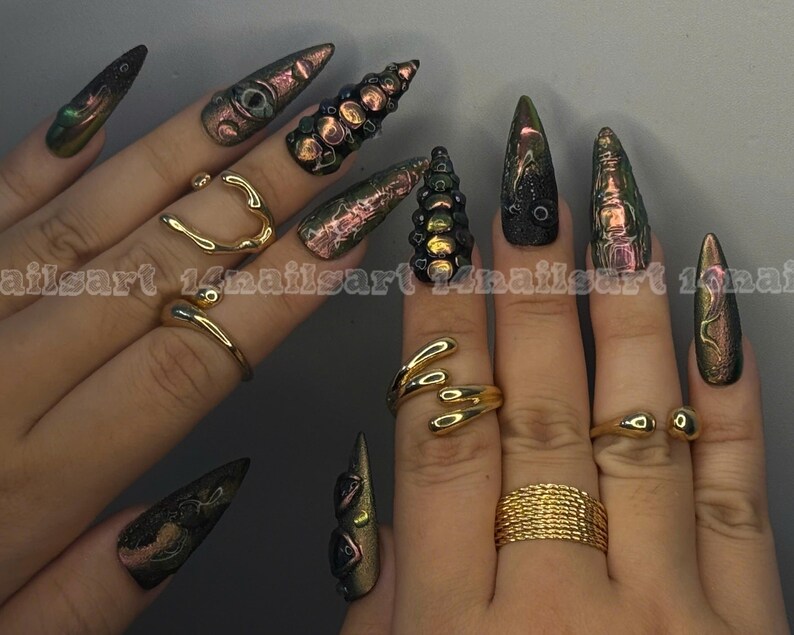 Peut inclure: Mains avec de longs ongles pointus iris&eacute;s verts, violets et dor&eacute;s. Les doigts sont orn&eacute;s de bagues dor&eacute;es de diff&eacute;rents mod&egrave;les, dont un c&oelig;ur, une forme enroul&eacute;e et une bande tress&eacute;e.